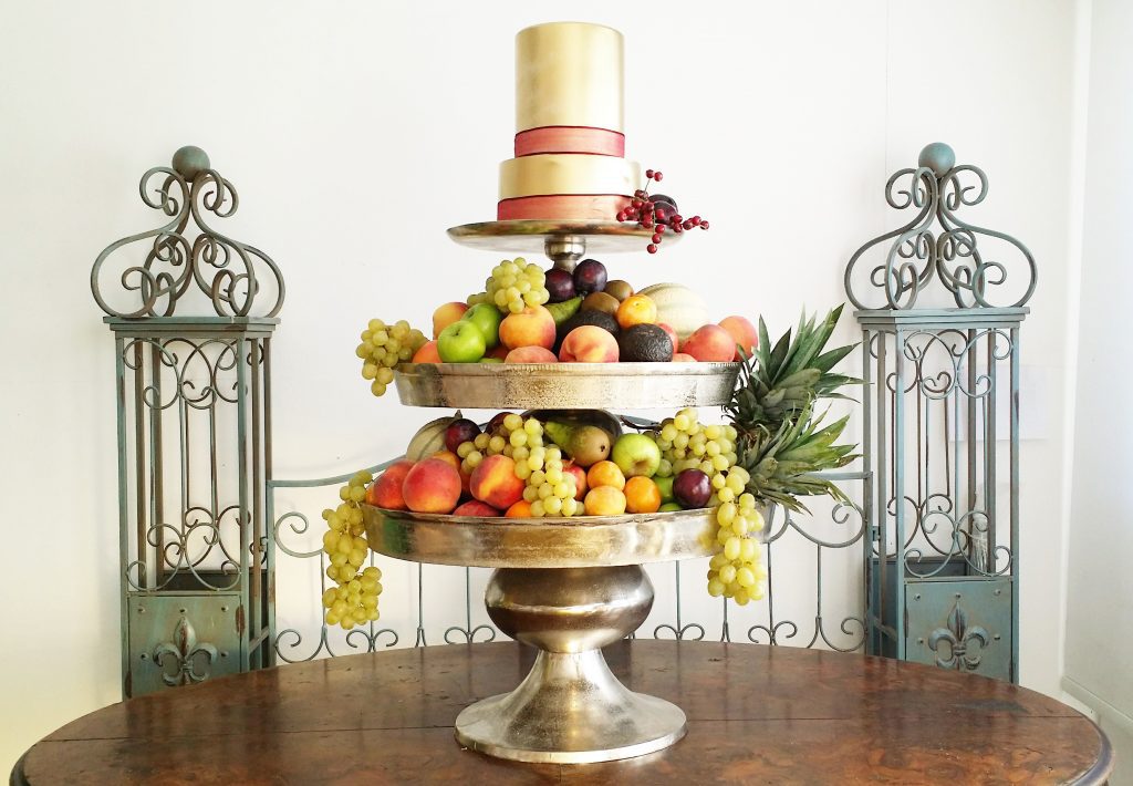 a grand layered three tiered silver cake stand. The bottom and middle layers are filled with grapes, peaches, apples and pears the top layer is a gold frosted two tier cake with a red ribbon around the bottom of each layer all available to hire