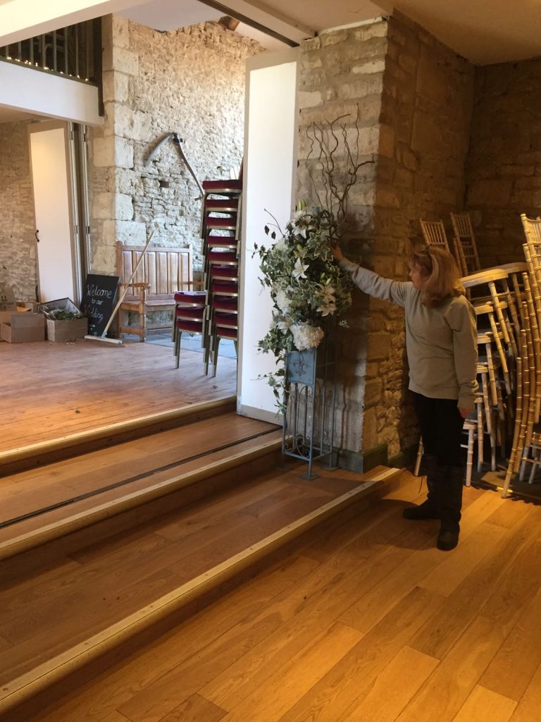 A member of our team finishing setting up tables at the great tythe barn in Tetbury with green hessian squares under a gold candelabra covered in a wreath of fake white roses and fresh season greenery surrounded but gold charger plates and a full place setting set up for hire