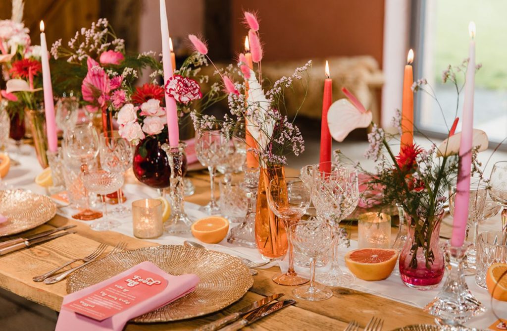 A wooden trestle table set up for a hen party with place settings of Gold glass charger plates and vintage cutlery with cut crystal wine water and champagne glasses. Down the centre of the table is cut crystal candlesticks with pink, orange and red candles, Red vases and glass bud vases filled with pink pampas and dried flowers.