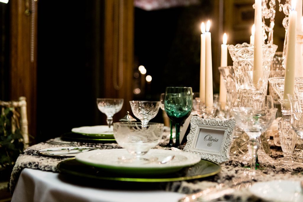 Close up of glass vintage candlesticks and a Fresh foliage wreath with ivy and hanging crystal chains centrepiece, with vintage glass vases and vintage glass candlestick and tealight holders and vintage wine, champagne and water glasses on a faux fur tablecloth to hire