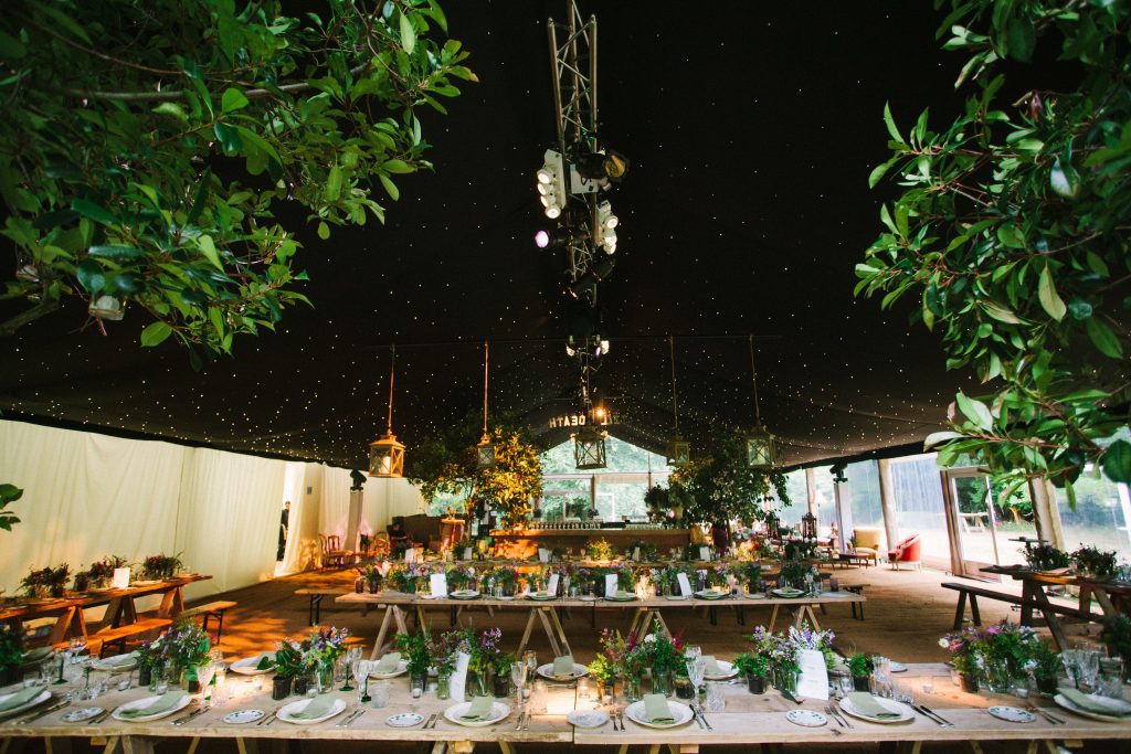 Marquee woodland wedding and event hire for a night under the stars, trestle tables with vases of wild flowers around a bar with hanging lanterns and bay trees and vintage furniture, sofas, armchairs and tables to create a cozy cornerfor hire