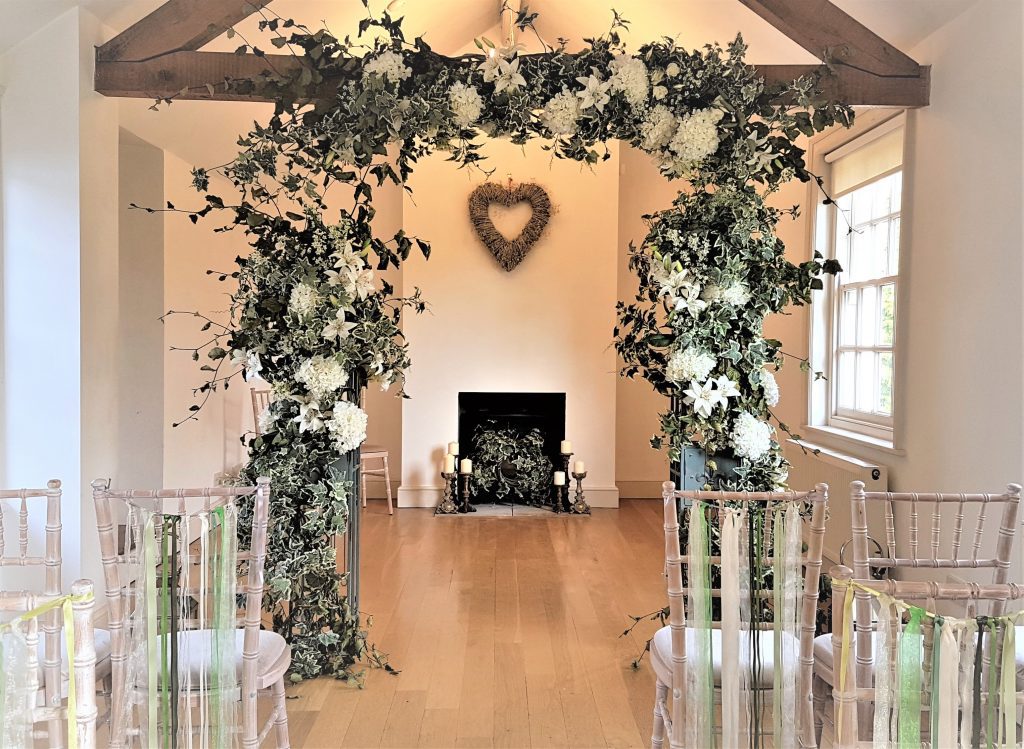 Enchanting wedding arch with faux or fake flowers in white and green, with fresh foliage and babies breathe with a rustic barn backdrop, perfect or an entrance way, available to hire for weddings and events