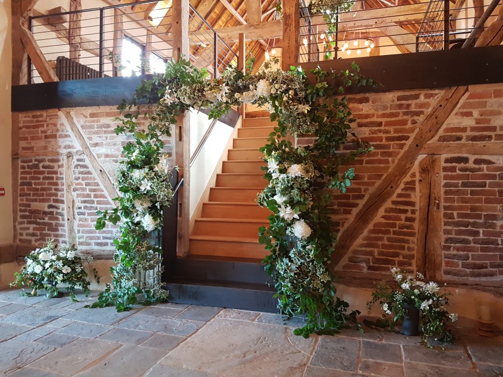 Enchanting wedding arch with faux or fake flowers in white and green, with fresh foliage and babies breathe with a rustic barn backdrop, perfect or an entrance way, available to hire for weddings and events