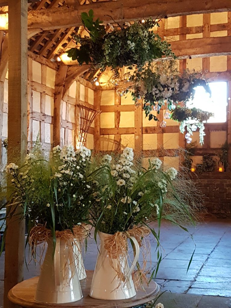 woodland wedding hanging wreath of white and green fake flowers and fresh foliage with rustic water jugs or water cans filled with wild flowers at Frampton Barn wedding venue available to hire for weddings and events