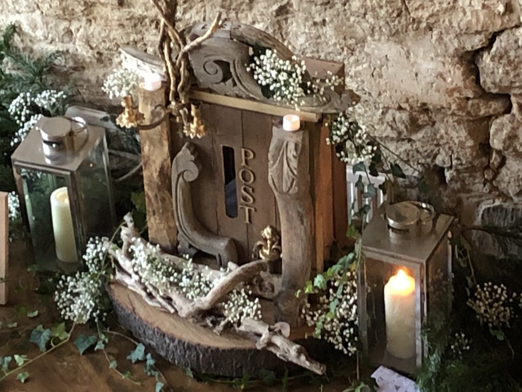 with a backdrop of Cotswold stone sits a Vintage rustic wooden crate post box with ivy and gypsophila, a silver lantern either side and winter pine branches, the guestbook and gift table available to hire at the Great Tythe Barn Tetbury