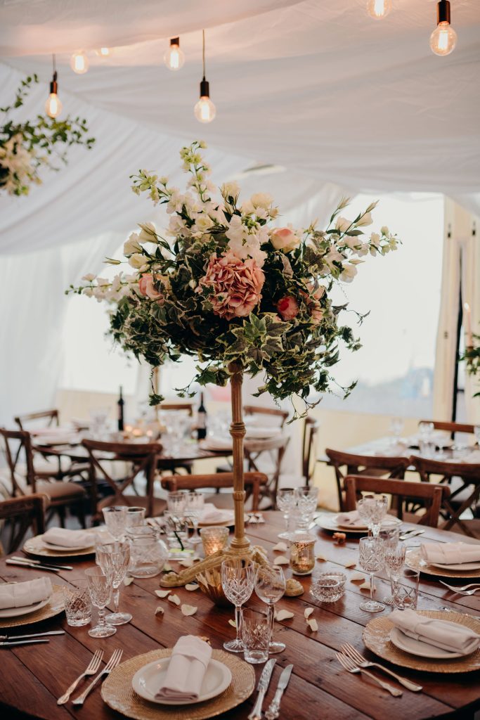 Floral wedding table decoration in a marquee a 10ft round table with a large floral arrangement with fake ivy, lisianthus, pink hydrangeas and gypsophila on a gold pedestal as a centrepiece surrounded by coloured and gold tealight holders and place settings of our gold charger plates and Vintage classic cutlery and glassware.