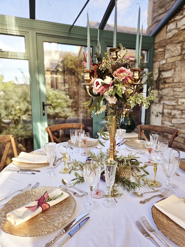 a centrepiece of a gold candelabra with a wreath of faux flowers in dusty rose  and fresh greenery including purple ribbons, around the base is fresh greenery and ivy for hire