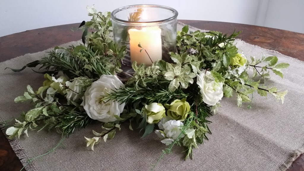 Small glass hurricane vase with a pillar candle and wreath of white roses and fresh greenery and lavender on a hessian table cloth to hire