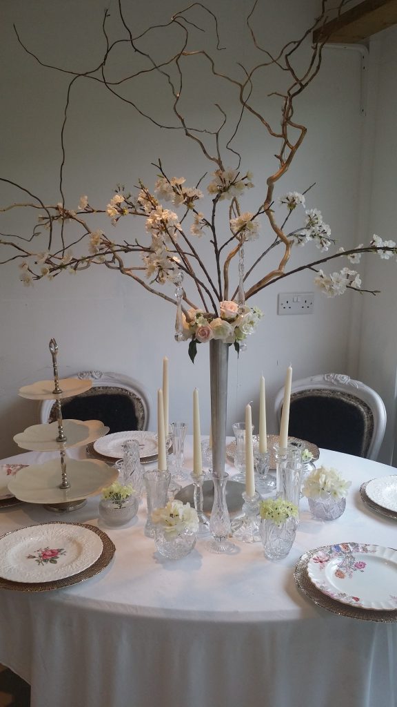 Wedding table of a slim silver vase with blossom branches, a wreath of roses, twisted willow and hanging crystals with cut crystal vintage vases and glass candlesticks around the base filled with pink hydrangeas and gypsophila and vintage crockery to hire
