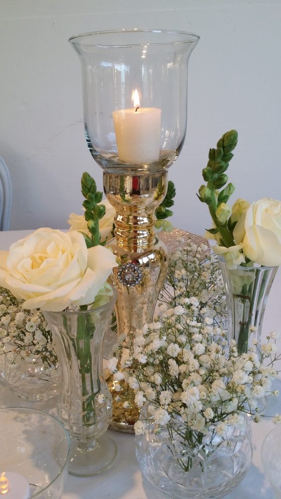 A Gold glass ornate tea light holder and trumpet vase surrounded by vintage cut crystal bud vases filled with gypsophila and white roses for a simple but elegant centrepiece