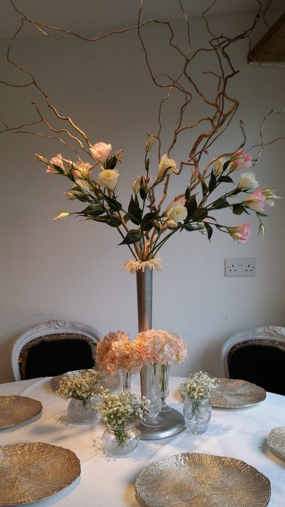 Wedding table of a slim silver vase with lisianthus, roses, twisted willow and pearls with cut crystal vintage vases around the base filled with pink hydrangeas and gypsophila and gold charger plates to hire