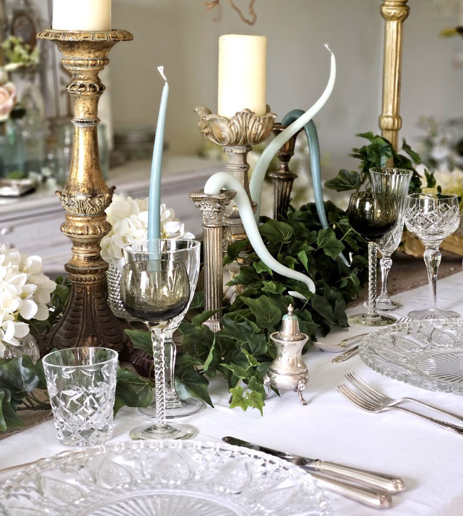 Centrepiece on a trestle table of Blenheim place gold pillar candlesticks with green ivy wrapped round the base and cut glass charger plates and vintage glasses for hire