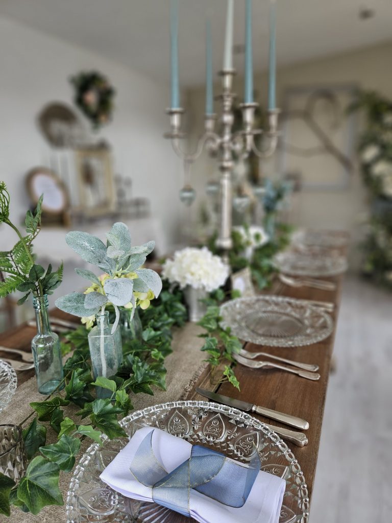 Centrepiece on a trestle table of Recycled glass bottles with single stem faux flowers and silver candelabras with blue glass baubles and teal candlesticks with green ivy wrapped round the base and cut glass charger plates and vintage glasses for hire