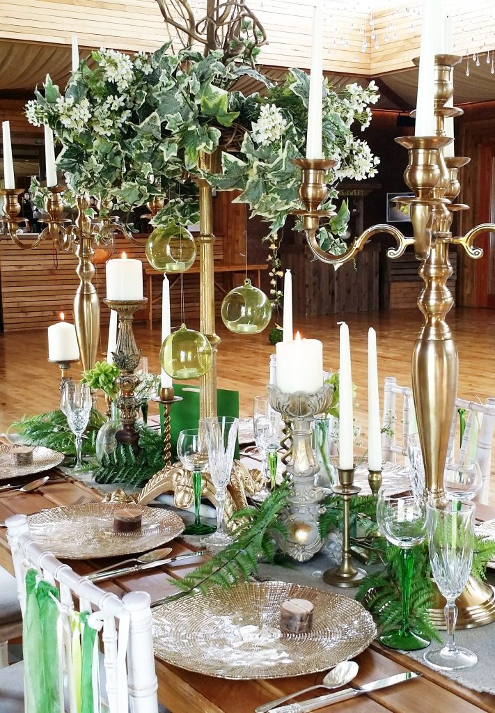 Gold candelabra and gold charger plates with a green ivy and twisted willow tall foliage table centrepiece, brass candle sticks and ornate pillar candle holders on a trestle table with a hessian runner to hire