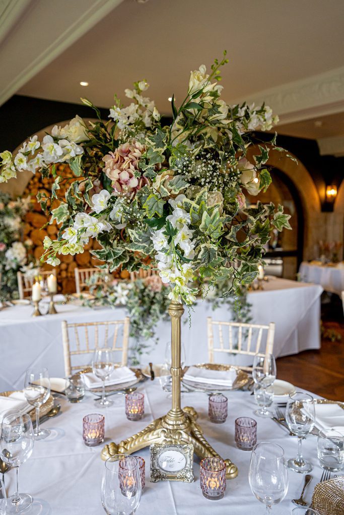 Floral wedding at the bear of Rodborough a 10ft round table with a large floral arrangement with fake ivy, lisianthus, pink hydrangeas and gypsophila on a gold pedestal as a centrepiece surrounded by coloured tealight holders and place settings of our gold charger plates and classic cutlery and glassware.