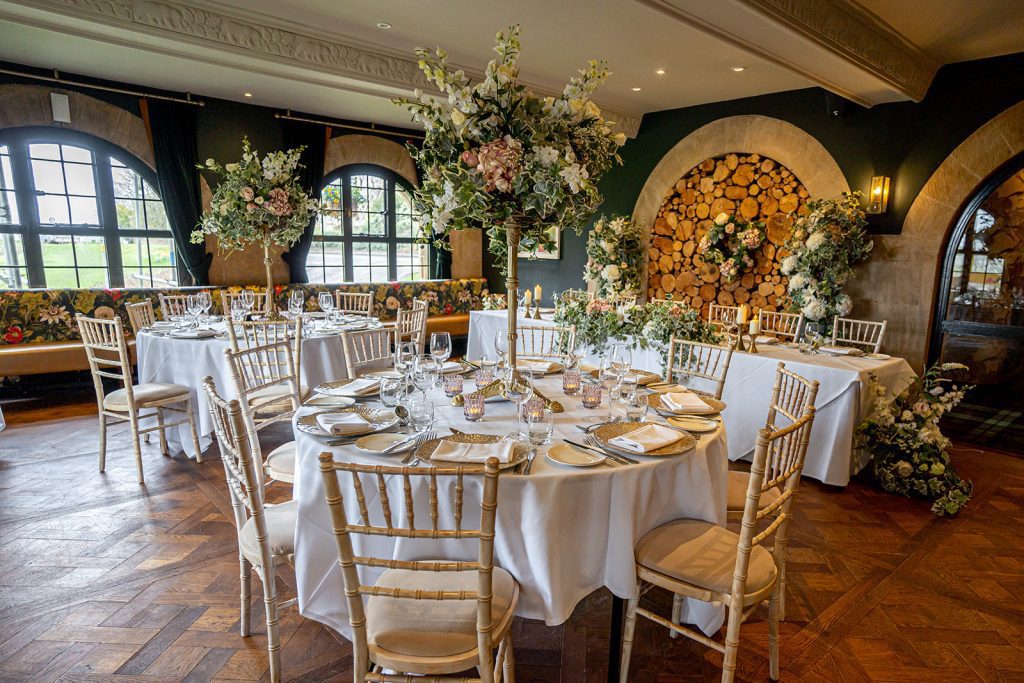 Floral wedding at the bear of Rodborough a trestle top table with a garland of ivy and pink and white roses with two pillars of faux flowers either side of the bride and grooms chairs. In the foreground two 10ft round tables with a large floral arrangement on gold pedestals as a centrepiece surrounded by coloured tealight holders and place settings of our gold charger plates and classic cutlery and glassware.