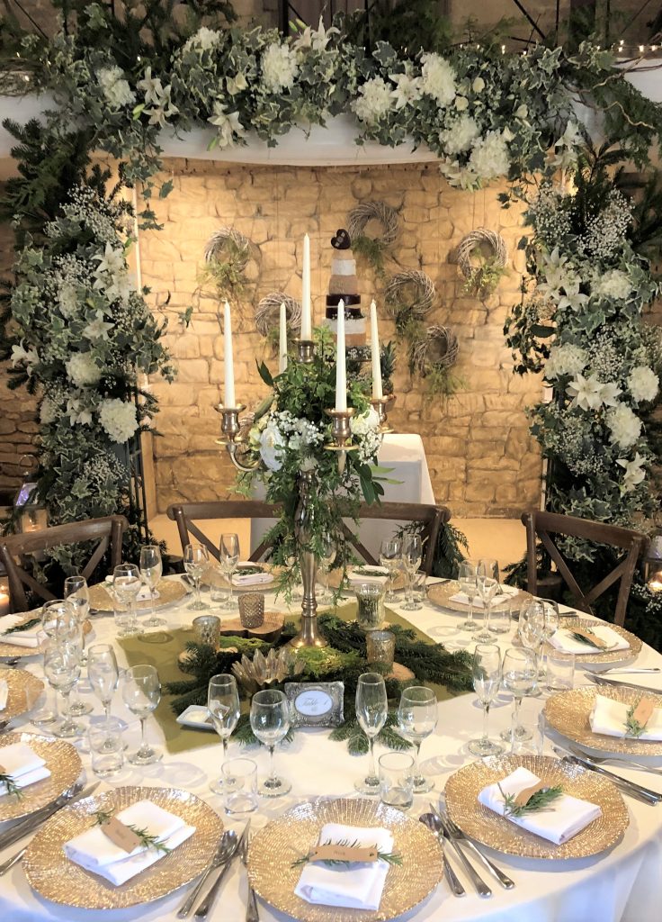 Gold candelabra, gold charger plates, and wooden slice with hessian table cloth and tealight holders in front of a white and green arch full of ivy and winter Christmas tree branches and a stacked cheese cake with rounds of Godminster cheeses including cheddar, stilton and brie for a woodland winter wedding available to hire