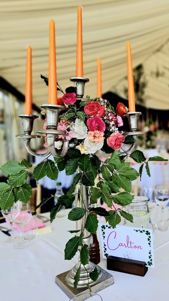 Centrepiece of a silver candelabra with a wreath of bright pink and orange roses and fresh beech trailing from the centre and orange candles for hire