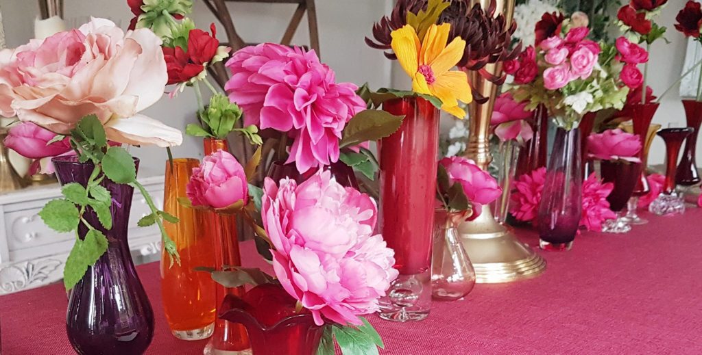 Magenta, pink and orange glass bud vases filled with bright pink dahlias, yellow cosmos and bunches of roses set on a magenta tablecloth and around a gold candelabra