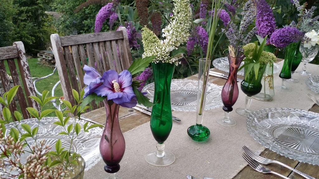 Purple and green glass bud vases with wild garden flowers on a hessian runner with cut glass charger plates and vintage cutlery table place settings