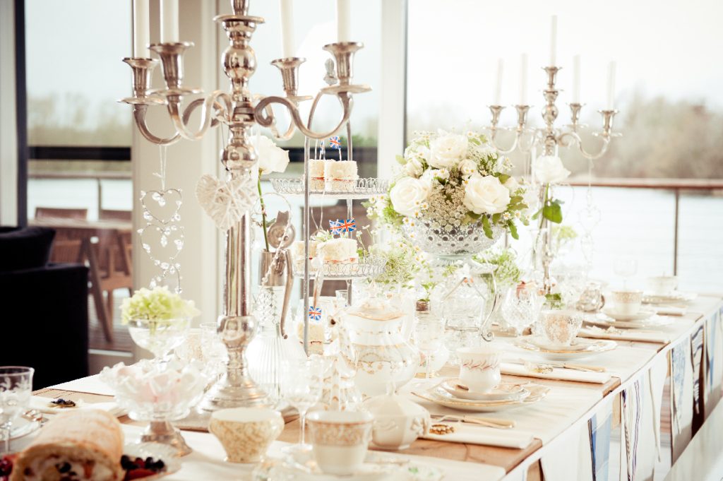 The lakes by Loo in the Cotswolds,  Vintage vases for a tea party table and Centrepiece is a large cut crystal punch bowl on a vintage glass pedestal cake stand, filled with a flower arrangement of large white roses, white carrot flowers,  gypsophila and fresh greenery. Either side is a three tier cupcake stand with mini Victoria sponges then there are two silver candelabras with hanging crystal hearts. the table place settings are made up with vintage crockery and tea cups and saucers and vintage sherry glasses, the table has a wrap of luxury quality white and blue bunting.