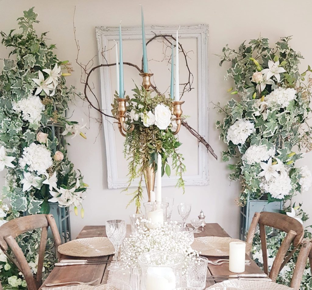 rustic wooden trestle table setup with gold charger plates, gold candelabra decorated with a wreath of white roses and fresh greenery, with place settings made up of vintage cut crystal wine glasses, tumblers and champagne saucers and cut crystal vintage vases with a backdrop of two pillars covered in faux white flowers and green ivy either side of a large vintage photo frame