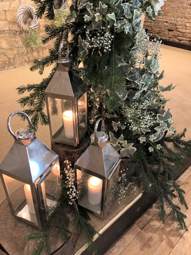 Three silver lanterns on wooden tree stumps in front of a frame of ivy, pine branches and gypsophila, for wedding ideas at great tythe barn, available to hire for weddings and events