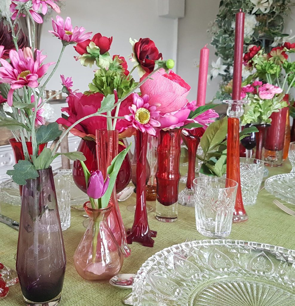 Magenta, red and aubergine glass bud vases filled with bright pink dahlias, red poppies and bunches of roses set on a eucalyptus green tablecloth with clear cut crystal charger plates, vintage glass tumblers and vintage cutlery