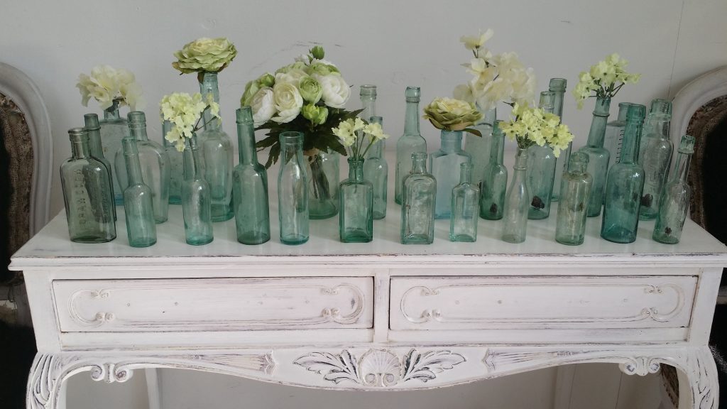 a large array of aqua green recycled glass bottles used for single stem flowers on a vintage side table