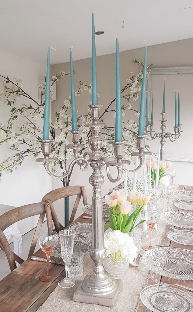 wooden trestle table made up for wedding inspiration with glass charger plate holding a white and navy vintage dinner plate and a smaller patterned glass side plate to the left. a hessian runner down the centre covered with vintage vases filled with gypsophila and cut glass candlesticks and two silver candelabras with blue candles with white blossom branches in the background