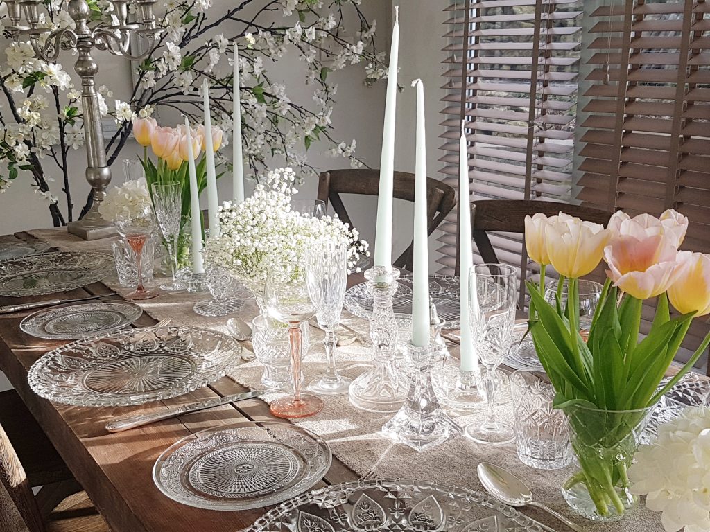 wooden trestle table made up for wedding inspiration with glass charger plate holding a white and navy vintage dinner plate and a smaller patterned glass side plate to the left. a hessian runner down the centre covered with vintage vases filled with gypsophila and cut glass candlesticks with white blossom branches in the background