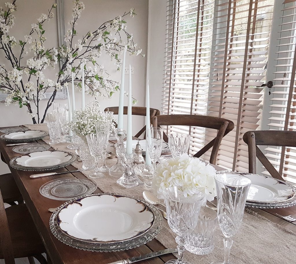 Glass charger plates holding white and navy vintage dinner plates and a smaller patterned glass side plate to the left. a hessian runner down the centre covered with vintage vases filled with gypsophila and cut glass candlesticks with white blossom branches in the background