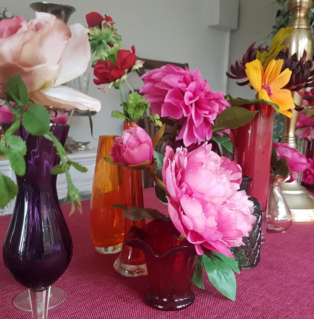 Magenta, pink, orange, red and taupe glass bud vases filled with bright pink dahlias, yellow cosmos and bunches of roses set on a magenta tablecloth and around a gold candelabra