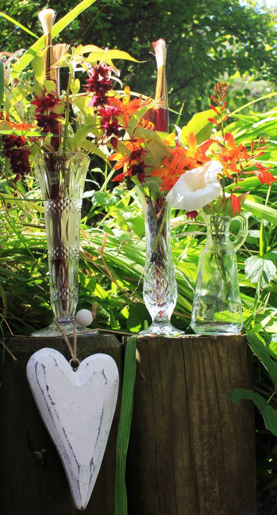 Three vintage crystal cut glass vases filled with wild garden flowers in aubergine, orange and yellow, with fresh greenery and a white wooden hanging heart in front of railway sleepers