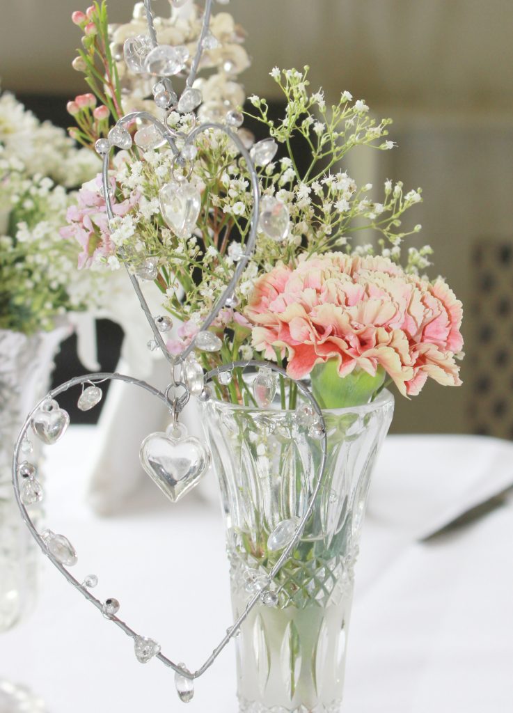 A vintage cut crystal glass bud vases filled with antique pink roses, pink hydrangea,  white daisies and gypsophila and sprigs of fresh eucalyptus and a silver hanging heart with crystals