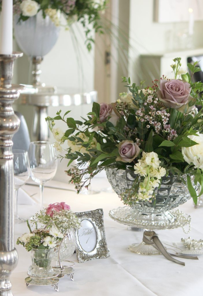 Vintage cut crystal punch bowl used for a flower arrangement on a glass cake stand, filled with roses and eucalyptus next to it a vintage glass and silver cruet set filled with wild flowers and a tall silver candelabra next to it