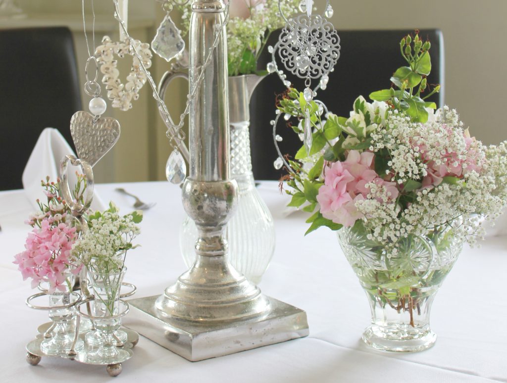 The base of a silver candelabra with hanging hearts surrounded by cut crystal vintage glass vases and a vintage silver cruet set and vintage cut crystal glass carafe filled with pink Hydrangeas, pink roses and Gypsophila.
