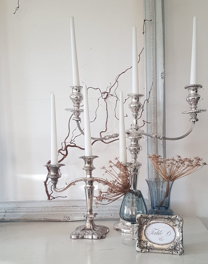 two Ornate silver three arm candelabras with blue vases infront and silver table number in the foreground with a grey vintage frame in the background